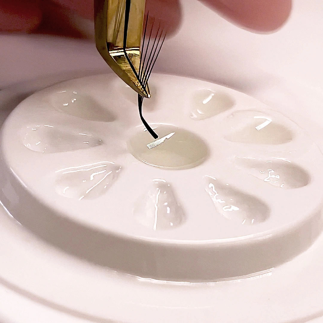 Macro shot of gold tweezers dipping the base of a volume eyelash extension fan into clear adhesive on a white glue palette.